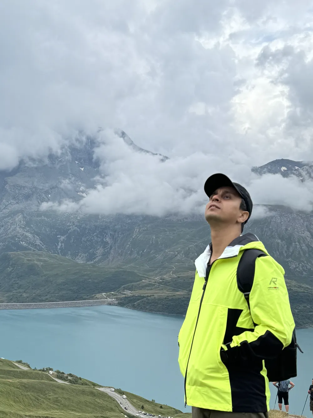 Abozar in a bright yellow jacket looking up at clouds over mountains and a turquoise lake