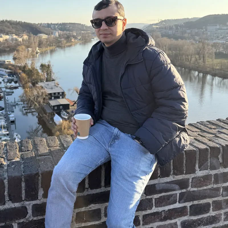 Abozar sitting on a brick wall with a coffee, the Vltava river below